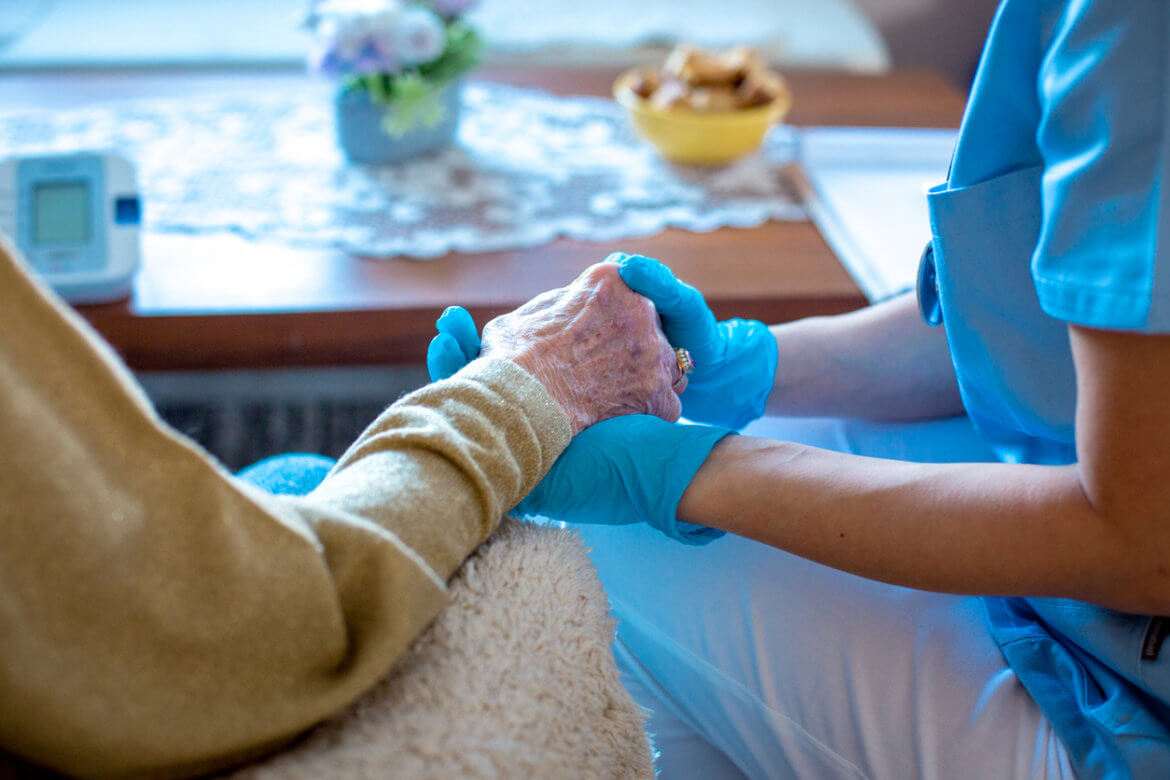 zClose-up shot of a health practitioner wearing blue gloves and holding the hand of a senior sitting beside them.