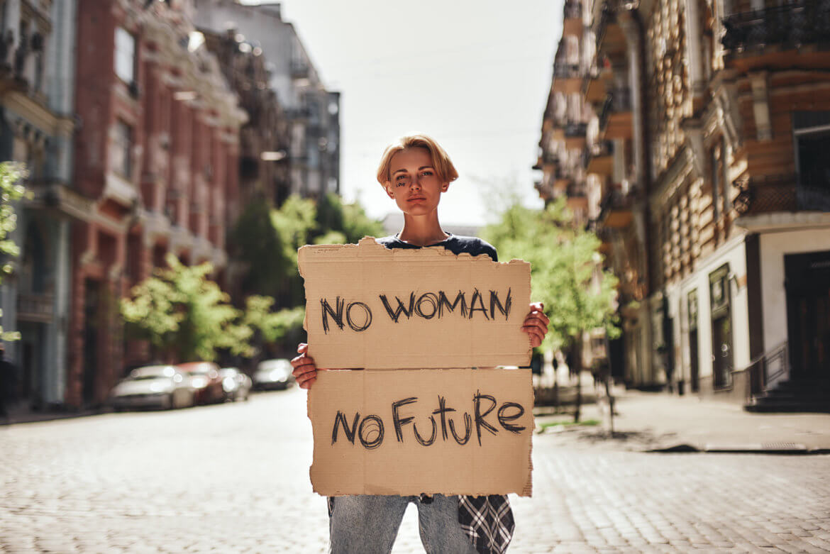 A woman stands in the middle of a cobblestone street holding a cardboard sign that says, “No woman, No future.”